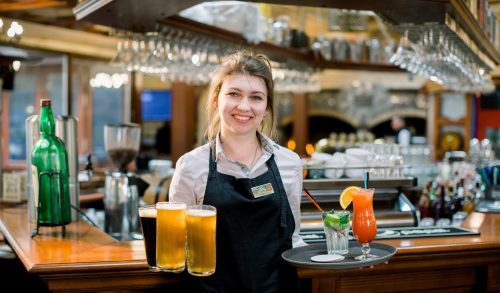 Smiling friendly waitress serving a pint of draft beer in a pub. Portrait of happy young woman serving beer in bar, looking at camera smiling